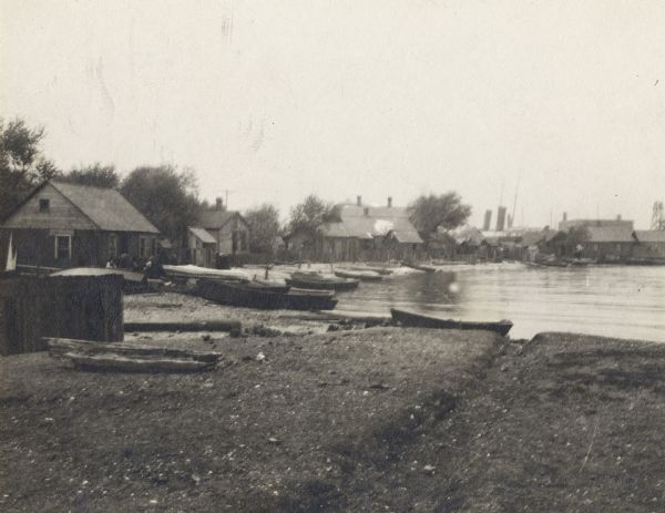 A fishing settlement. Small buildings and boats line a bay, with sand and a piece of driftwood in the foreground.