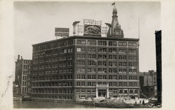 Building from across the river, with construction workers working nearby. Signs for the Boston Store and Diamond tires are on the roof, as well as lettered signs in various windows.  A tall building with a clock tower rises in the background.