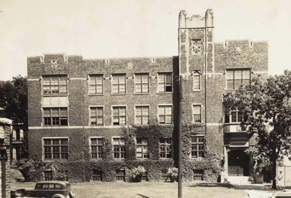 Four-story building, with vines growing on the exterior.  A woman stands near the entrance on the right. Above the doorway a sign says "Dentistry". A car is parked in front on the left.