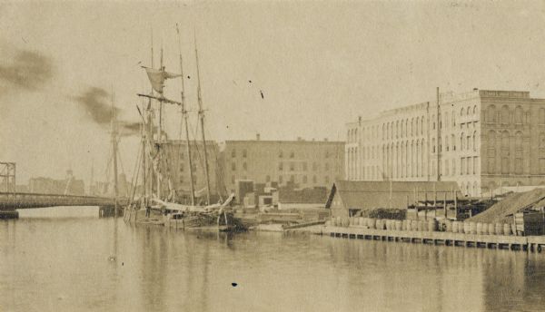 North of Huron Street Bridge looking toward point near Grand Avenue crossing.  A ship with several masts is docked on the river, and several large buildings are along the shoreline. A line of barrels are on a pier near a small wood building.