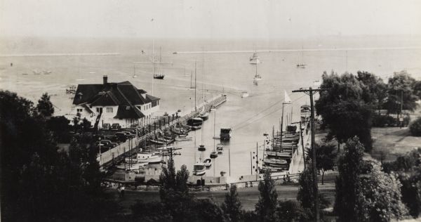 View from hill of the yacht club, with Lake Michigan behind. In the distance is the jetty, and the road along the shoreline is N. Lincoln Memorial Drive. There are boats moored near the club, and other boats out on the water.