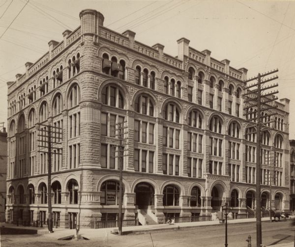 Also the Northwestern Mutual Life Insurance Company building. Large building on the corner of National Avenue, with a sign for the National Exchange Bank painted on the window. A young boy is standing on the corner, and a young girl is walking with an adult and a baby carriage along the sidewalk. There is also a horse-drawn buggy parked on the street, which is lined with electric and telephone poles. Located at the corner of Broadway and Michigan Street.