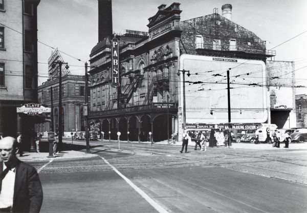 Corner of Wells and Water Streets. A large blank billboard is on the side of the theatre. Across the road on the left is a sign for Blatz beer. A car wash sign is on the back right of the theatre by the parking lot. Pedestrians are on the sidewalk and crossing the street.
