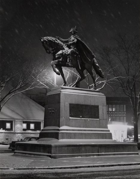 In Washington park, Washington Park library in the background.  Nighttime view, with snow falling. The statue of Frederick Von Steuben on horseback presides over the street. A Revolutionary War hero, the German-born Von Steuben (1730-1794) served with George Washington. His statue, sculpted by Swiss-born artist J. Otto Schweizer, was dedicated in 1921 and stands as an impressive gateway to the park.