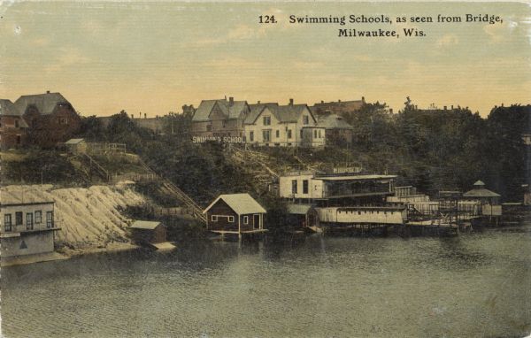 Elevated view from bridge looking over at a hill leading down to bath houses and fenced-in swimming areas for beginners. Establishment of Wm. Bechstein. Caption reads: "Swimming Schools as seen from Bridge, Milwaukee, Wis."