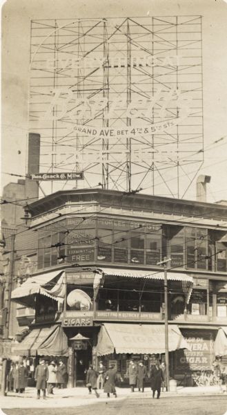 A corner store with a large electric sign on top. Men are on the sidewalk in front of the store, and a horse-drawn vehicle and bicycles are on the right.
