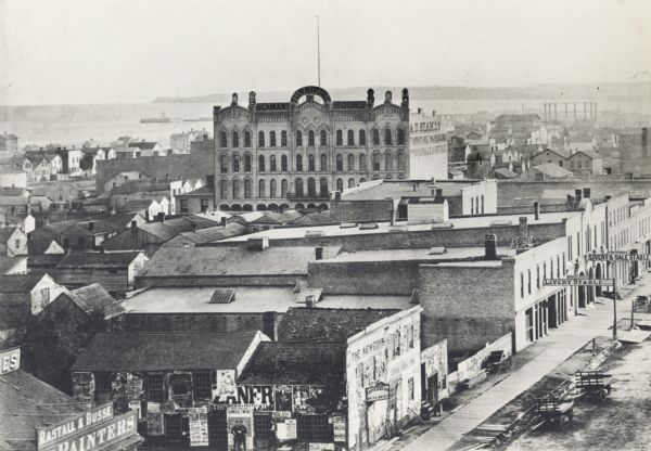 Elevated view over rooftops looking towards Lake Michigan shoreline. Seaman's Furniture and Rooms building is surrounded by other commercial buildings. There is a sign for "Livery and Sale, Stable" on the lower right. Carriages and carts are parked along the street.