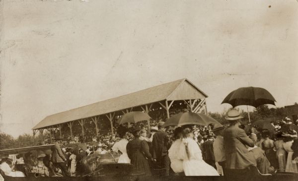 Crowd at fairground, with people watching from horse-drawn vehicles in the foreground, more spectators in the grandstands with umbrellas in the background.