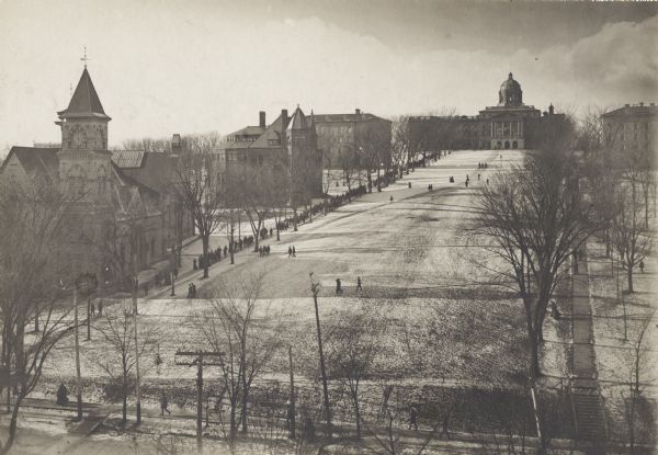 Elevated view of Bascom Hill and lower campus vicinity from the roof of the Wisconsin Historical Society, which was then under construction. A large group of pedestrians are walking on the path on the right, and there is snow on the ground. Bascom Hall (formerly Main Hall) is at the top of the hill. Music Hall with the clock tower is on the left.