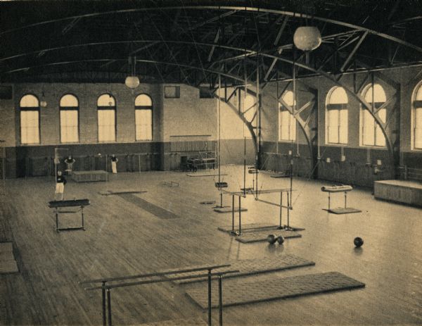 Elevated view of interior of gymnasium at University of Wisconsin-Madison. Three men stand in the room, facing the viewer. Various gymnastic and weight equipment is placed throughout the gym.