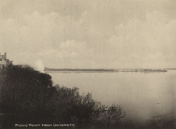 Picnic Point from shoreline of Lake Mendota at the University of Wisconsin-Madison campus. There is a university building on the hill on the left, and a pier with a boat in the foreground.