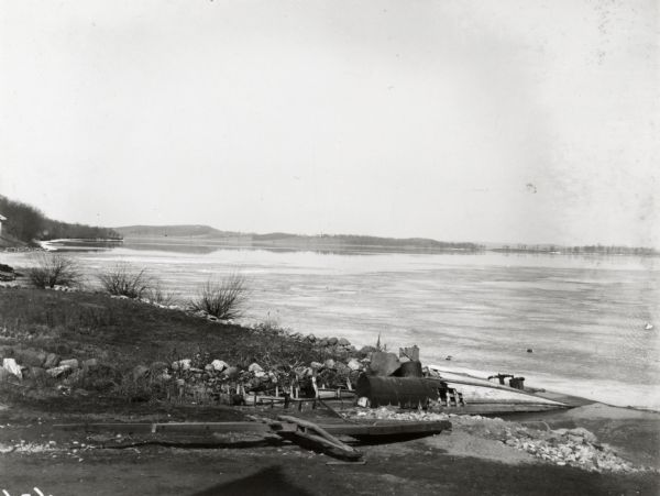 View from Chi Psi porch on the University of Wisconsin-Madison campus. Picnic Point is stretching towards the right in the far background. There appears to be a boat hull and boiler in the foreground and snow and ice along the shoreline.