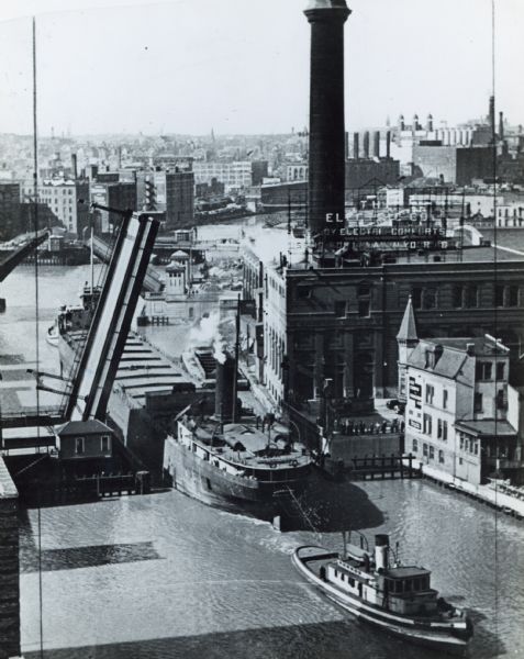 Elevated view of a tugboat pulling a lake steamer on the Milwaukee River under an open drawbridge near the Electric Company.