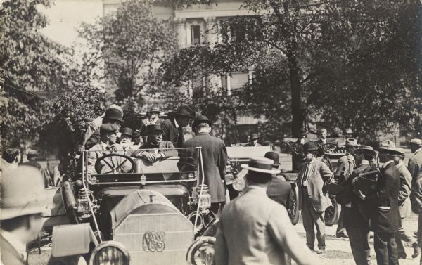 A group of men are sitting in a car among a crowd of people. Standing on the right are two photographers. Theodore Roosevelt is in the backseat of the car on the right side.