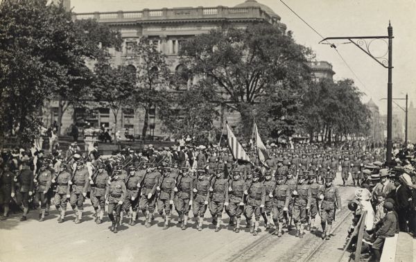 Slightly elevated view of soldiers marching in a parade down a street. Spectators fill the sidewalks.