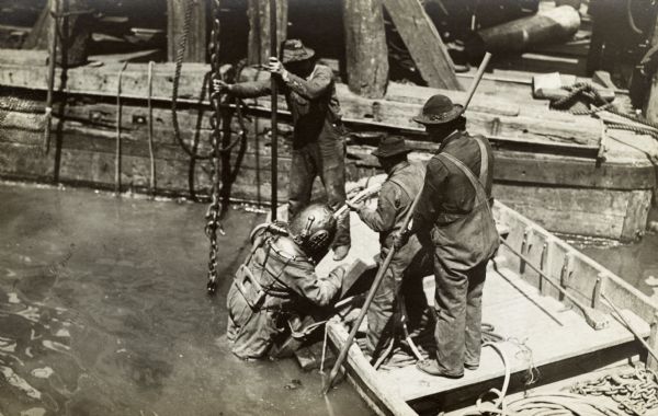 Man in diving suit being helped out of the water by three men in a rowboat.