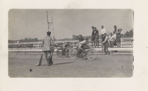 Two motorcyclists are racing by while several men are watching from behind a railing. One man standing on the track in the foreground is holding a flag.