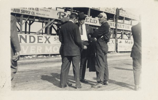 The man in the goggles on the right is Teddy Tetzlaff; possibly at the Vanderbilt Cup Race. The other men are unidentified. In the background is the grandstand area with large advertisements.