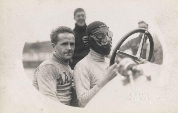 Two men sitting in a racecar, with the driver of the car, possibly Harry Endicott, wearing a leather mask with goggles. Two men are standing in the background.