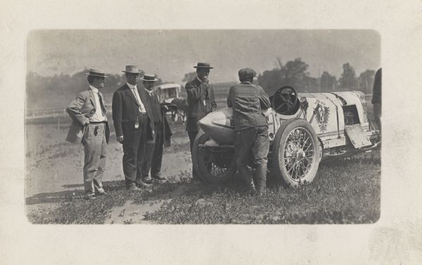 Several men are standing around a white car, with one leaning on the back end. A panel near the front of the car is open, exposing part of the engine. A European heraldic symbol is on the side of the car. The car is a 1909 Blitzen Benz, the driver of the car was Bob Burman. The car raced in the 1911 Indy 500.