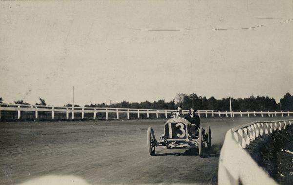 A two-passenger car with the number 13 on the front is going around the bend on a dirt race track. The guard rail fence is in the foreground.