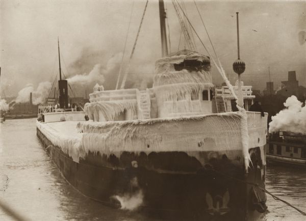 A tugboat in the river is covered in snow and ice. Two men are at the front of the ship on the right wearing hats and heavy coats.
