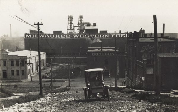 A car climbs up a dirt and gravel road in an industrial area. The Milwaukee-Western Fuel Co. building is in the background, as well as a building with a sign that reads: "Cooperage".