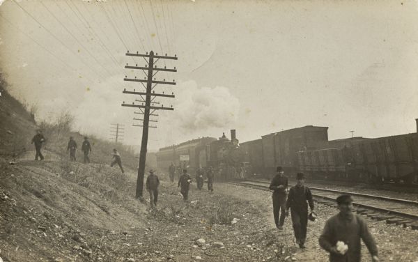 A group of men carrying lunch boxes are walking away from a steam engine train, which is near another train and power lines.