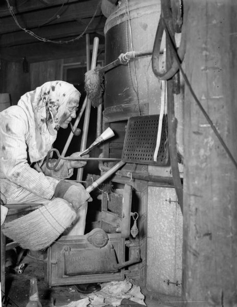 J. Robert Taylor cleaning a furnace. He has a broom and a small brush in hand, a pipe in his mouth, and a dirty rag placed on his head. He is reading a document he is holding in his hand.