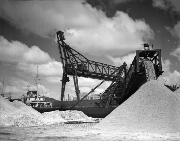 Piles of sand are on the ground, and more sand is pouring off a large dredge. The dredge is mounted on a ship. A building in the background has a water tower on the roof.
