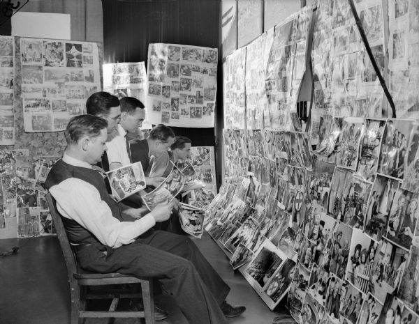 Newspaper workers perusing hundreds of photographs. There are three men holding some of the photographs and the rest are mounted on the walls around them.