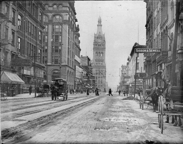 Pedestrians, bicyclists, and horse-drawn carriages on a commercial street. City Hall with its clock tower is in the far background. A sign for Sisson & Sewell is on the right.
