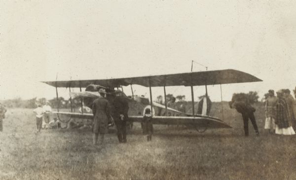 A crowd of adults and children looks over a JN-4HG gunnery trainer airplane / biplane which has landed in a field.  Airplane has a black cat symbol in a circle and the number 44214 or 44216 on the side. Titled in the album "The First Aeroplane in Madison". "The feature of the flight was the delivery of a letter to Postmaster Devine from Pres. William F. Brooks of the Minneapolis Aero Club.  This is the first aerial letter ever received in Madison and the first aerial postage stamp ever received here.  The flyers are returning from a 3,000 mile run and have been making exhibition flights at state fairs."  The Capital Times 9-4-1918. One of five airplane images taken over a two day period.