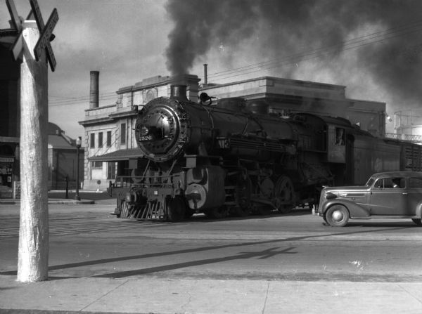 A train is shown at the intersection of Williamson, S. Blair, and E. Wilson Streets in front of the Chicago and Northwestern Railroad Depot.