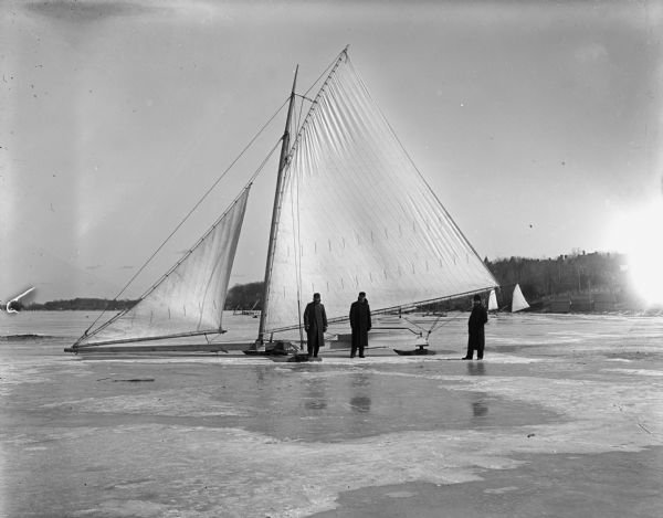 Three men stand near a large iceboat owned by Dutton and Wheeler on Lake Mendota.