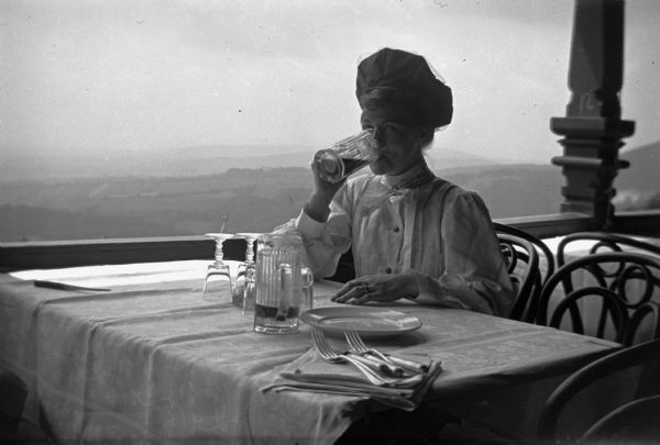 Mary Smith, wife of Dr. Joseph Smith, drinks a beer on a veranda or balcony while on sojourn in Austria. Mr. and Mrs. Smith stayed in Austria for eight months while on a special study at the University of Vienna.