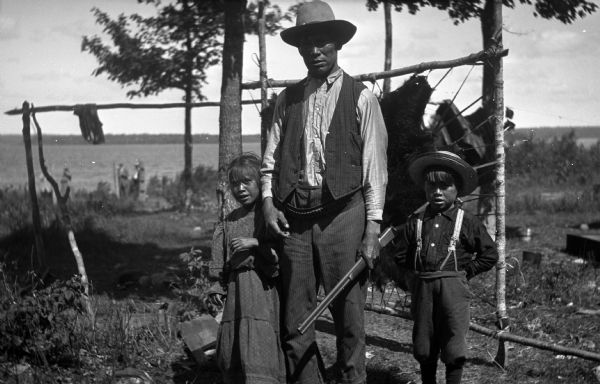 View towards a man standing with two children, a boy wearing a hat and suspenders, and a girl wearing a dress. They are standing in front of a bear hide stretched on poles, with a lake in the background. The man is holding a rifle and is wearing a hat, vest, shirt, and trousers. The man and children are Lac Vieux Desert Chippewa Indians.