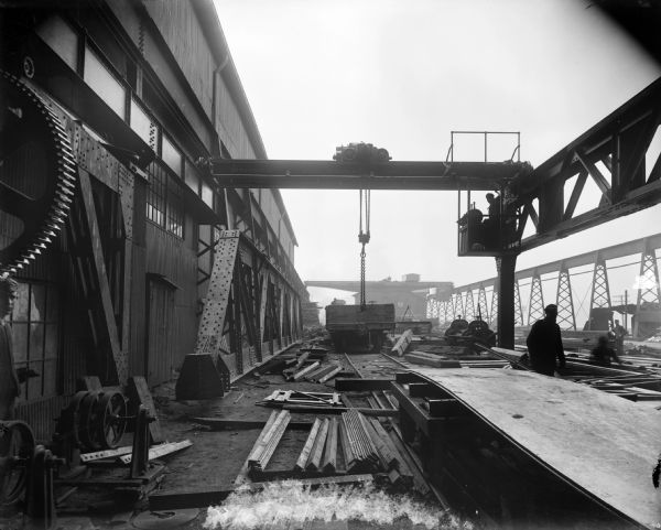 An interior 5-ton cab-operated Pawling and Harnischfeger crane running along railroad siding to unload scrap and steel for manufacture. The stencil on the crane reads "Pawling and Harnischfeger Builders, Milwaukee, Wis. Capacity 10,000 Lbs." There is a man in the cab of the crane as well as men working on the ground. The crane is attached to a building on the left, and supported on the right by a crane runaway.