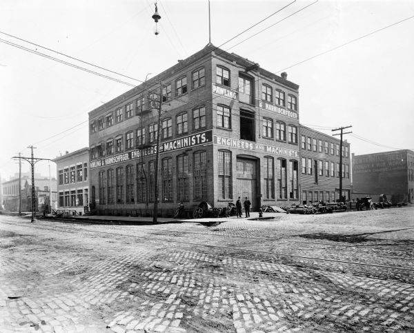Exterior view of one of the first Pawling & Harnischfeger machine shops. The building was built in 1886 on the corner of South First Street and East Oregon Street. Painted on both sides of the building are the words "Pawling & Harnischfeger, Engineers and Machinists." There are five men standing in front of and to the left of the building. Painted on the side of the building to the right of the machine shop are the words "Centennial Bell Foundry & Iron Works, Gardiner Campbell and Sons."