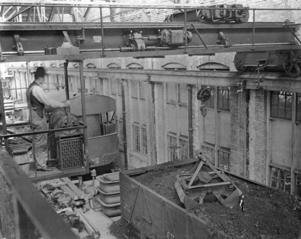 View near crane operator looking down on coal car near exterior of building. Pawling & Harnischfeger type "O" bridge crane with a type "G" trolley in a foundry operation unloading a coal car with a grab bucket unit. The crane is stamped "Patented, Pat. No's., 647436, 697393." The man is operating the dial controls of the cab.