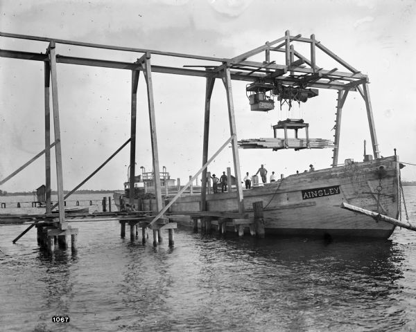 Pawling & Harnischfeger 5-ton monorail hoist with a lumber handling unit, possibly in Pensacola, Florida. The monorail track extends over the dock to permit loading barges and boats with lumber. The text on the front of the ship reads "Ainsley" and there's a man in the cab, men on board, and men on the dock.