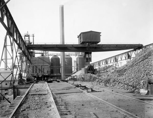 A Pawling & Harnischfeger type "AN" hoist with a grab bucket and an enclosed trolley over railroad tracks with a train car underneath it. The crane is stamped "Pawling & Harnischfeger." A man is looking out of the cab, another man stands on the left, and chimneys and storage tanks are in the background.