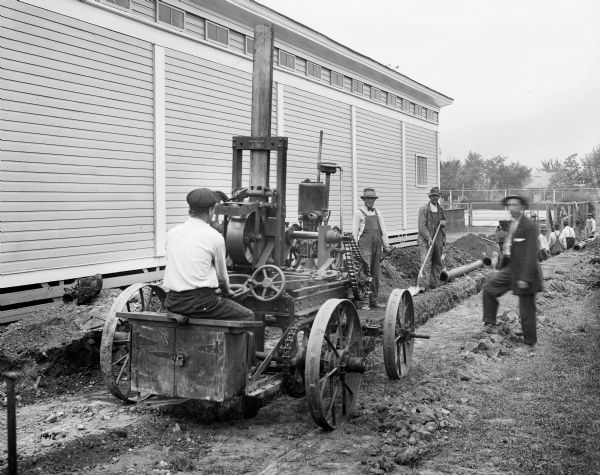 Gas-Powered Concrete Tamper/Breaker Man operating a gas-powered concrete tamper/breaker that is being used to consolidate concrete over ditches after laying water or sewage main pipes. There is a building on the left, a few men stand near the tamper in the foreground, and in the background men are working down in the ditch.
