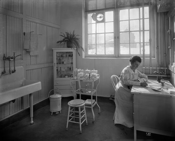 The first aid department at a Pawling & Harnischfeger plant. A woman sitting at the desk working on the "daily report first aid room." On the left is a sink, in the corner there is a medicine cabinet, and behind her a window looks to the outdoors towards another building.