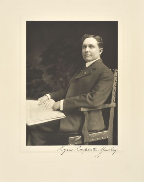 Three-quarter length seated studio portrait in front of a painted backdrop of Cyrus Carpenter Yawkey, Wausau manufacturer. He is sitting in a chair at a table and holds something in his hand.