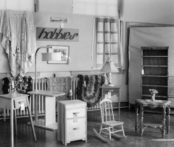 A hobby show with quilts hanging from the wall. Rugs are draped over radiators. There are also bedside tables, lamps, a bookshelf, and a rocking chair. A sign on the wall reads "hobbies."