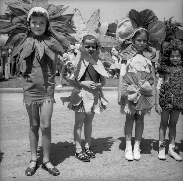 Four girls standing in a row dressed as flowers, probably as part of a Fourth of July "Garden Queen" pageant. Each of the girls has a number around her arm.