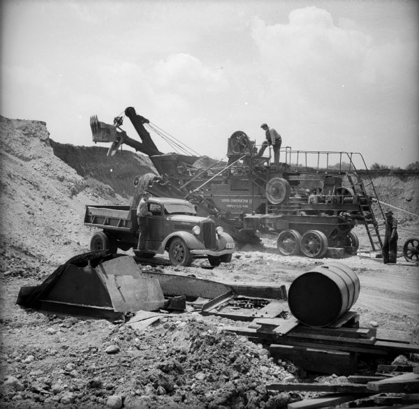 A man is on a walkway on top of a mechanical conveyor belt machine that is loading sand and gravel into the bed of a truck. A steam shovel is visible in the background. The conveyor machine has the words "Burns Construction CC / Minneapolis Minn." written on its side. A man stands at the back of the conveyor machine and another man is on the running board of the truck.