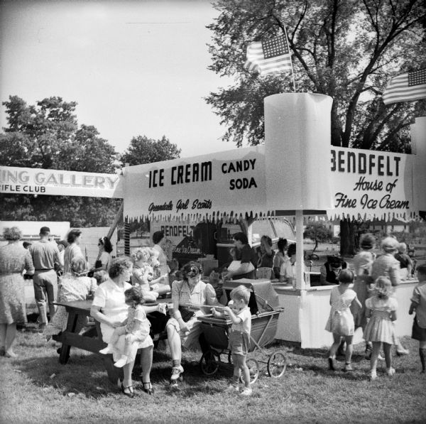 A scene from the Greendale Civilian Defense Rally. Mothers and children sit at a picnic table outside of the "Bendfelt House of Fine Ice Cream" stand operated by the "Greendale Girl Scouts." The ice cream stand is situated next to the "Shooting Gallery Rifle Club."