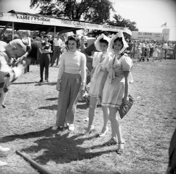 A clown entertains three women at a Civilian Defense Rally, two of whom are wearing bonnets and matching dresses. Booths behind the women read "Variety Store,""Prize Cakes & Coffee," the "Country Store," and "Ice Cream."